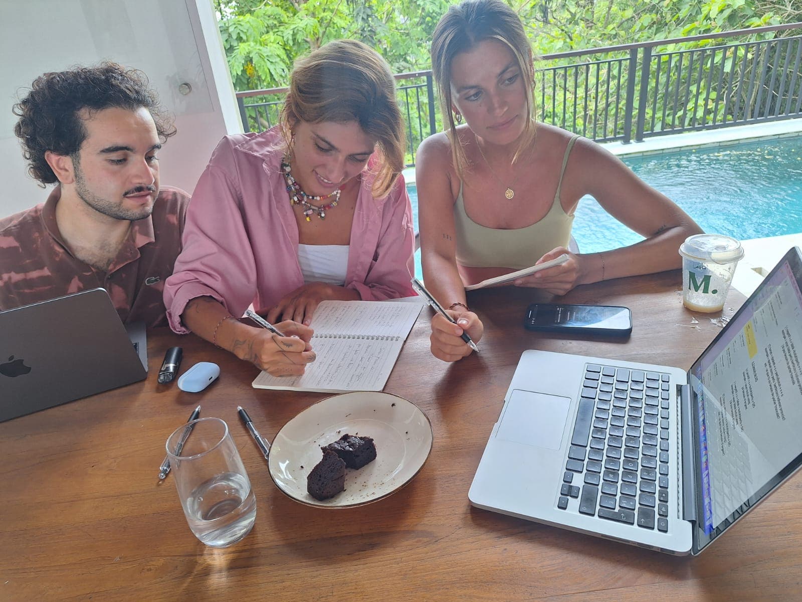 Trois jeunes travaillent sur une terrasse avec ordinateurs et cahiers près d'une piscine.
