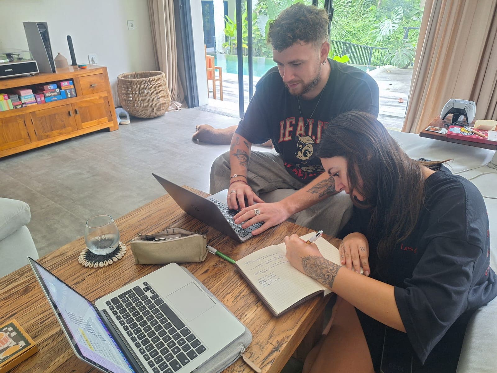 Un homme et une femme tatoués travaillent sur une table basse avec ordinateurs et carnet.