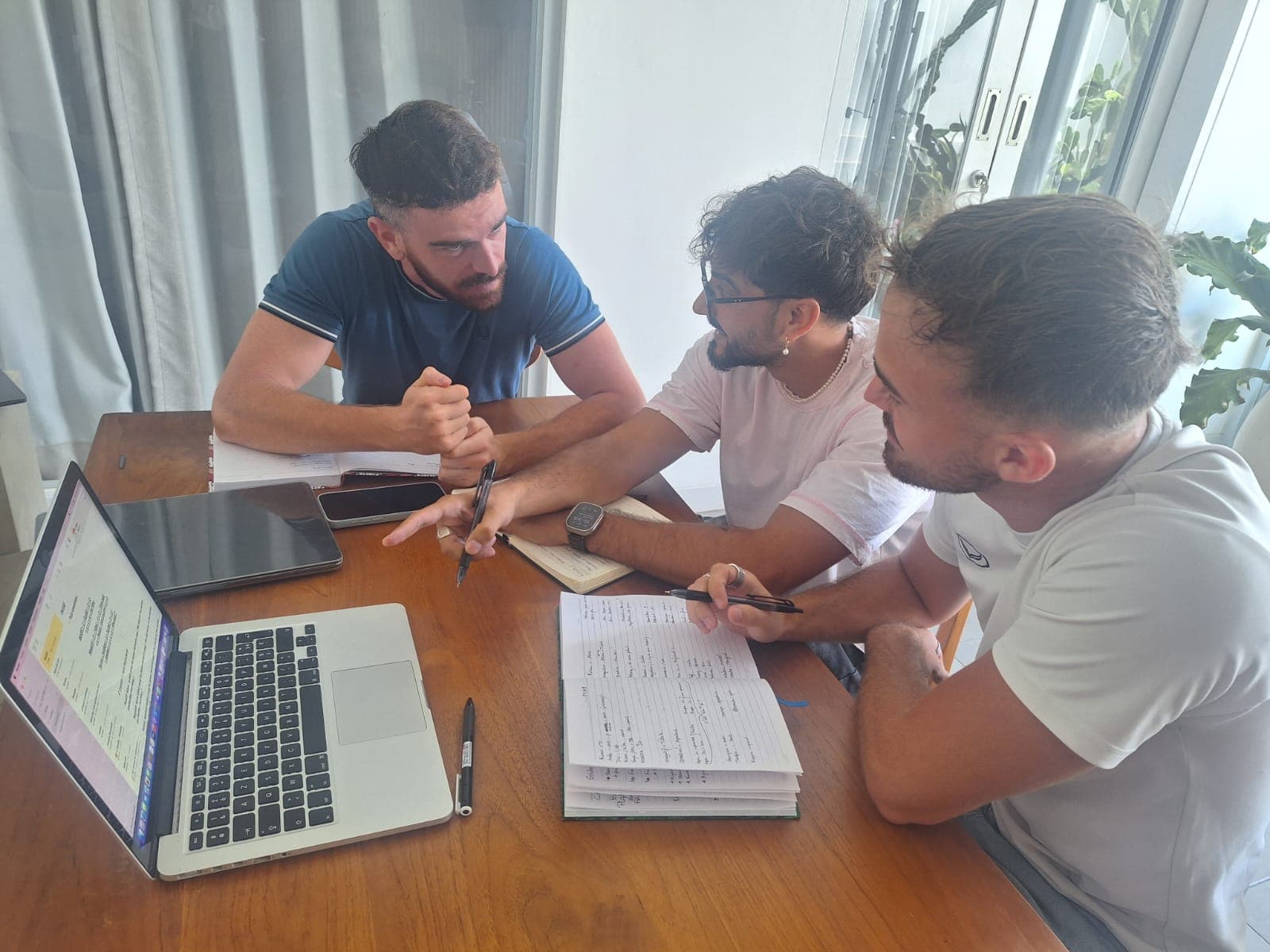 Trois jeunes hommes travaillent ensemble autour d'une table avec un ordinateur et des cahiers ouverts.
