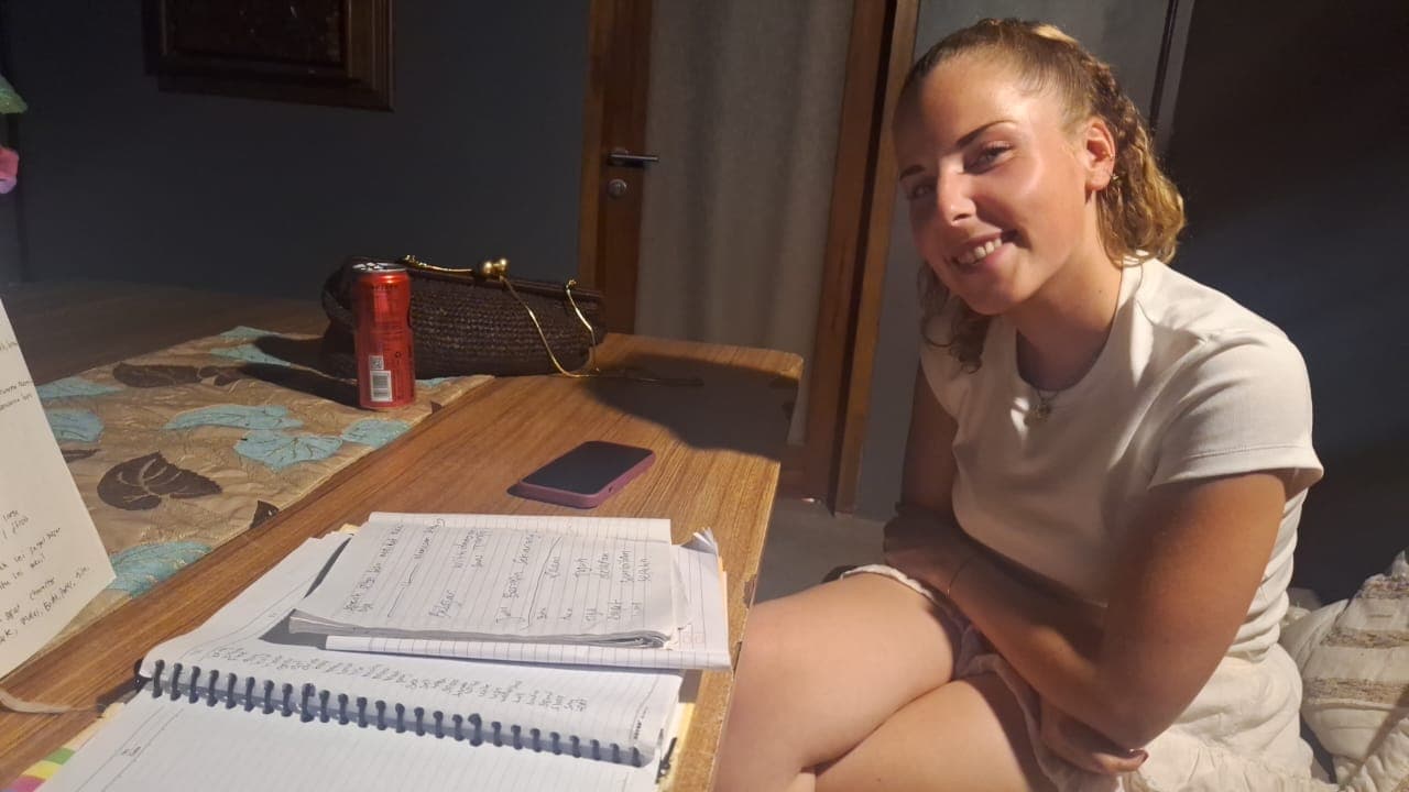Jeune femme souriante assise devant des cahiers ouverts, un téléphone et une canette rouge.