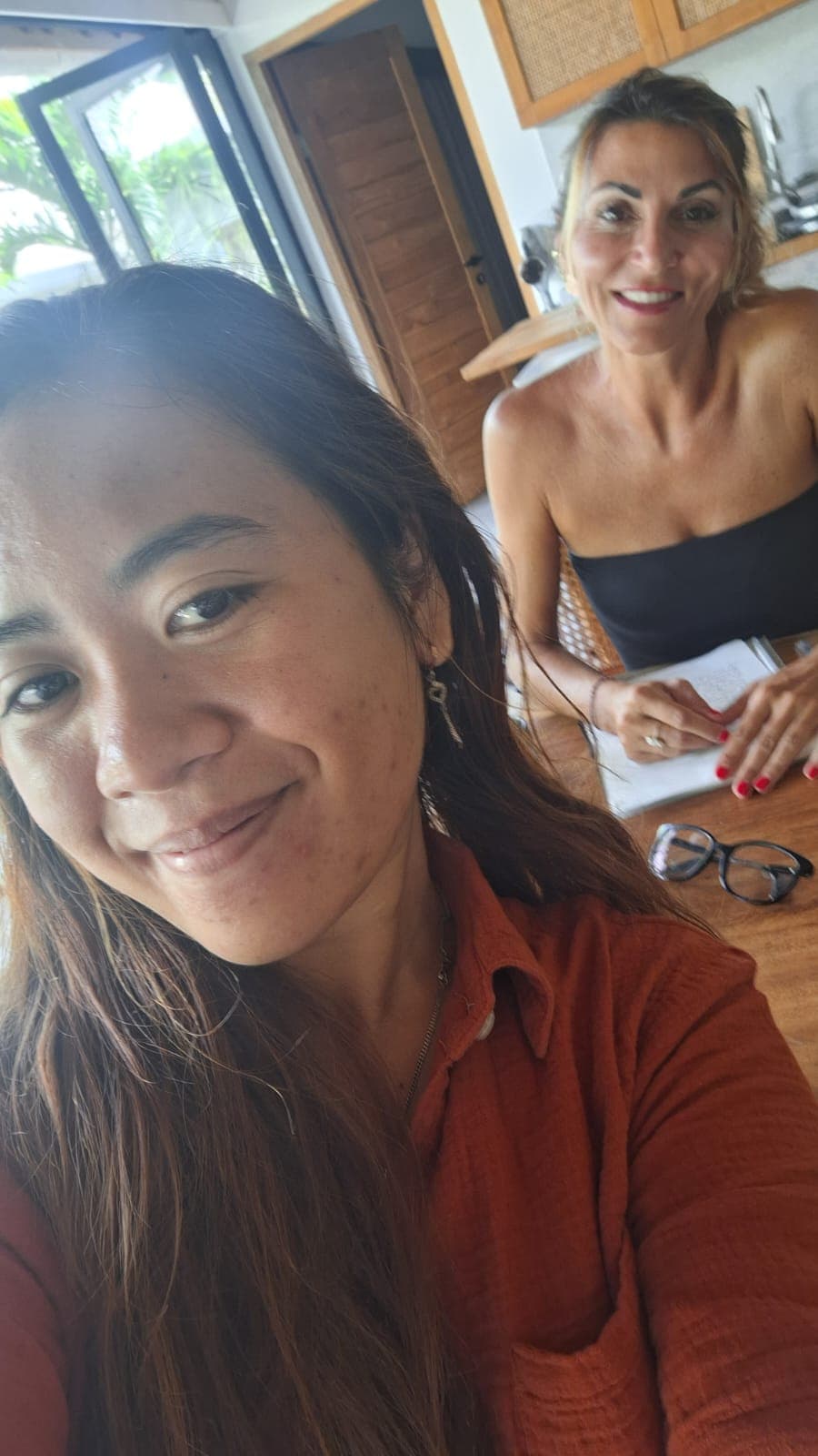 Deux femmes souriantes, l'une prenant un selfie en chemise orange, l'autre assise à une table.