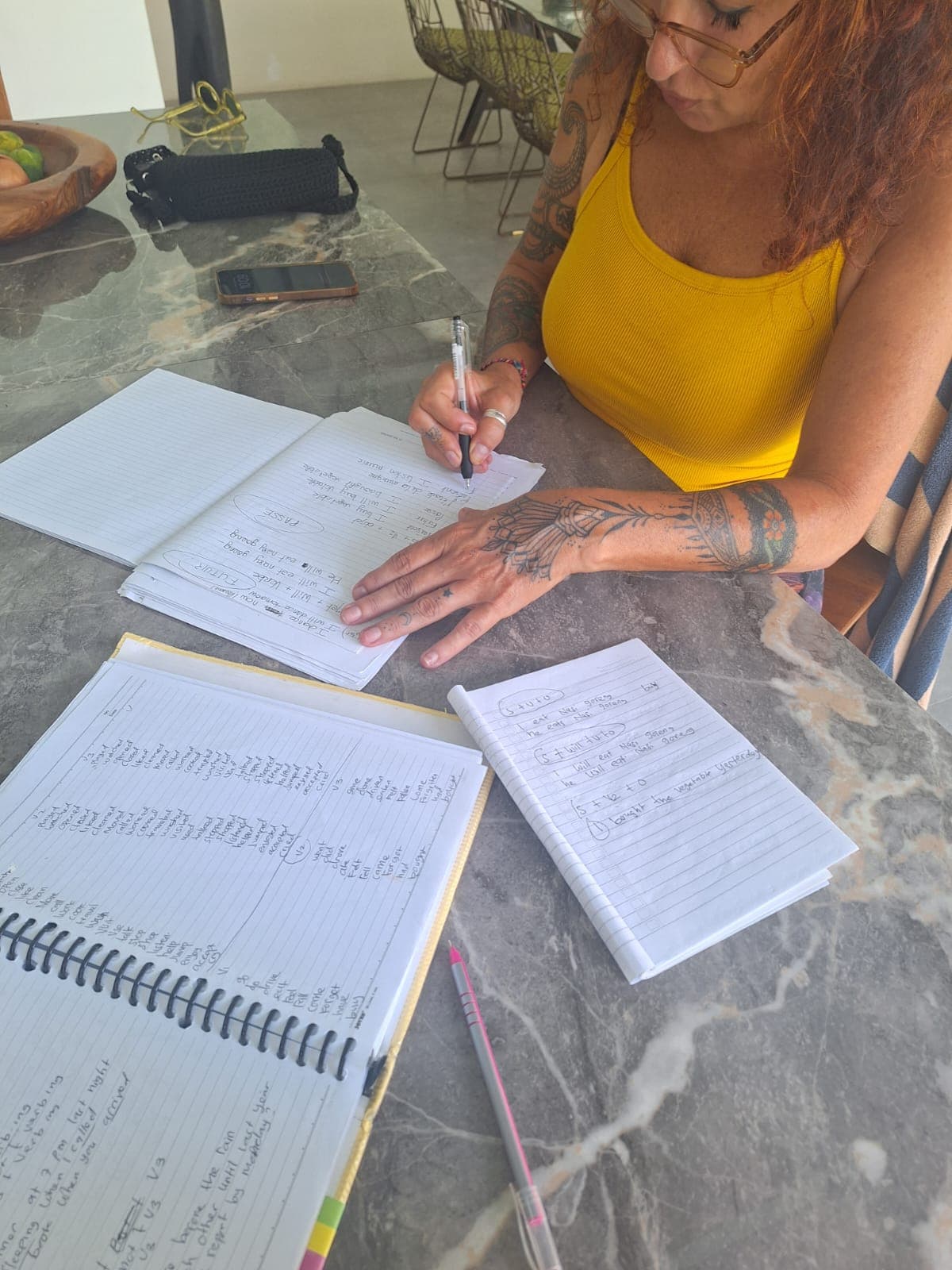Une femme tatouée en débardeur jaune écrit dans un cahier sur une table marbrée.