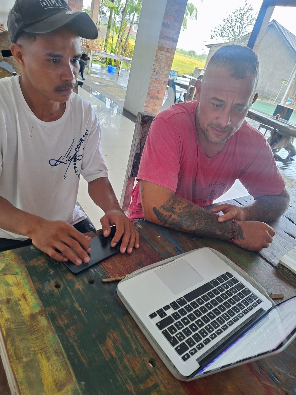 Deux hommes assis à une table rustique consultent un ordinateur portable et un smartphone.